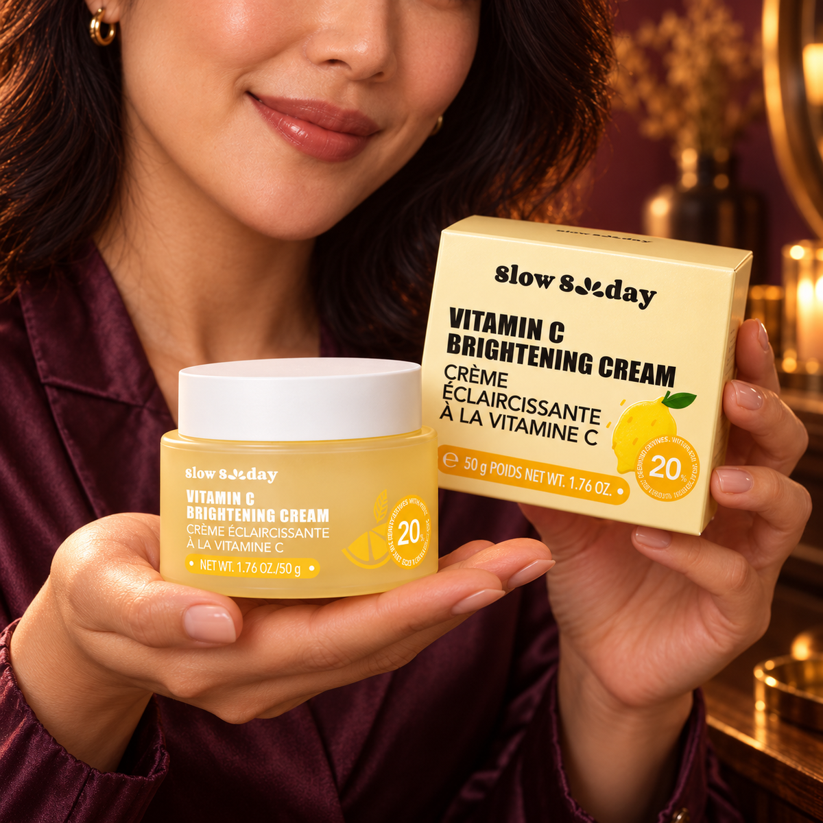Close hand-held package detail of the Slow Sunday Vitamin C Brightening Cream 50 g closed yellow jar and retail carton, presented by the same woman